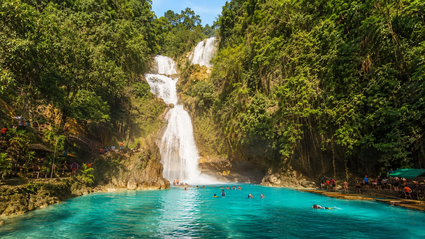 Kawasan Falls main waterfall Badian Cebu Philippines — three-tiered cascading waterfall with vivid turquoise-blue mineral spring water and canyoneers swimming in the pool at the base