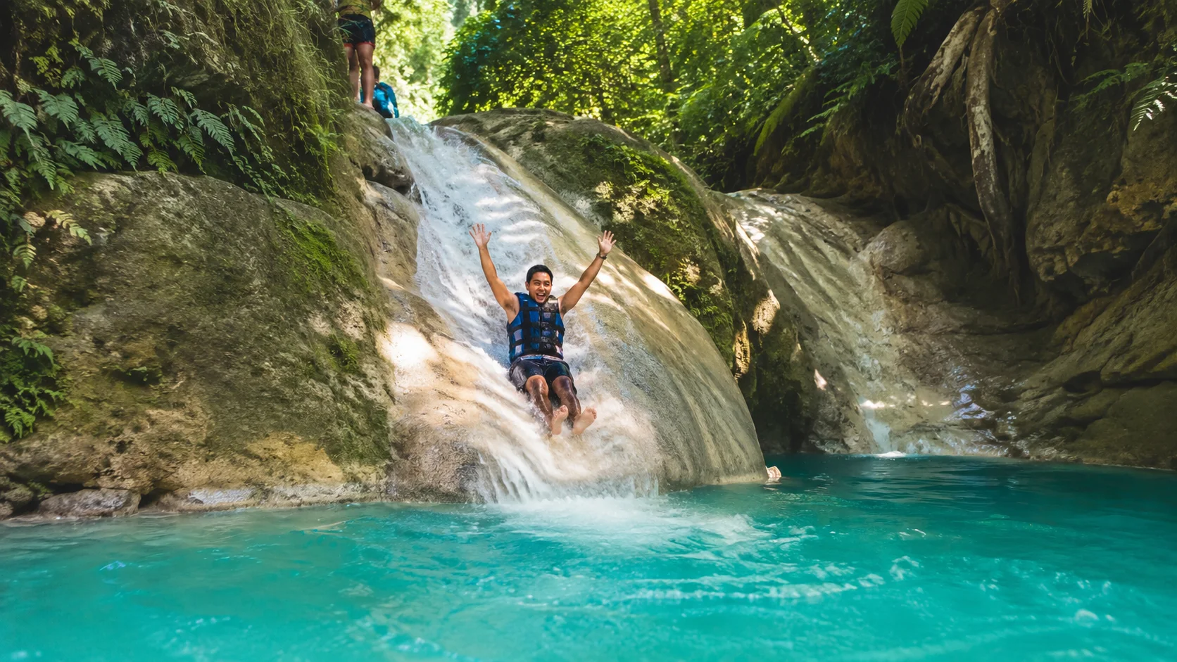 Natural rock slide at Kawasan Falls canyoneering Badian Cebu — tourist sliding feet-first down smooth limestone water chute into turquoise pool below