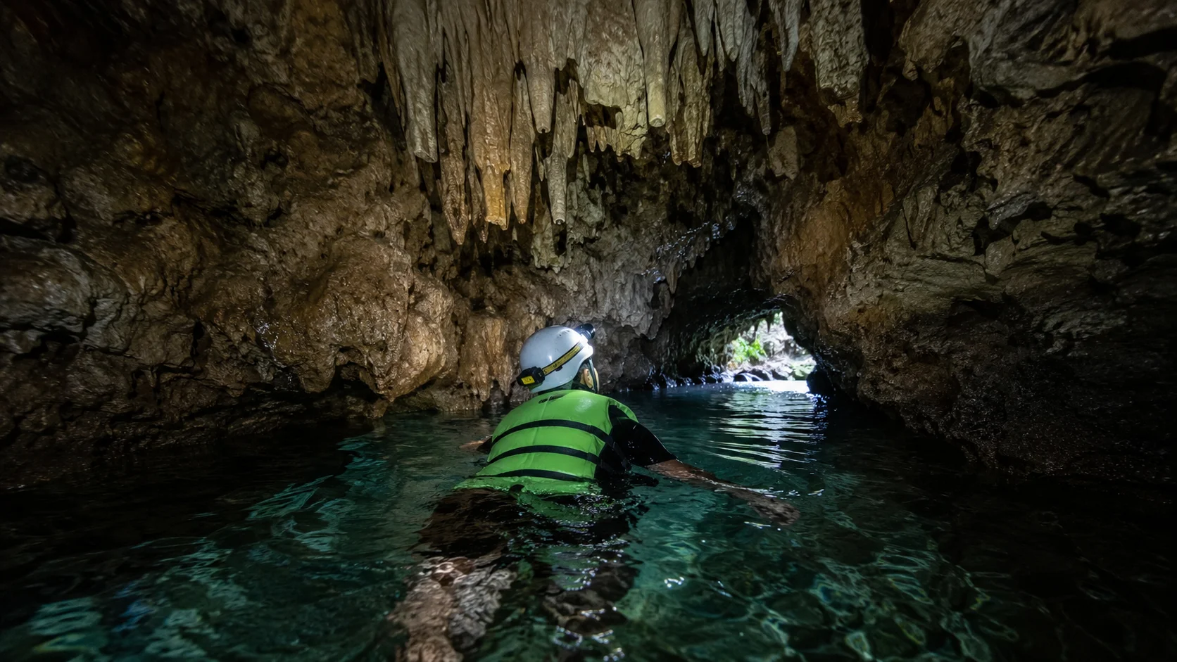 Stalactite cave swim at Kawasan Falls canyoneering Cebu — tourist swimming through dark limestone river cave with stalactites overhead, lit by headlamp in Badian Cebu Philippines